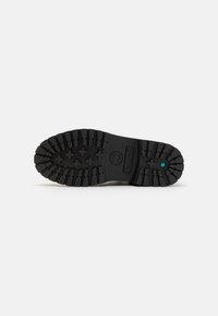 Suela de bota de goma negra con surcos profundos y elementos de tracción. Presenta un logo circular y un acento azul cerca del talón. Superficie texturizada.