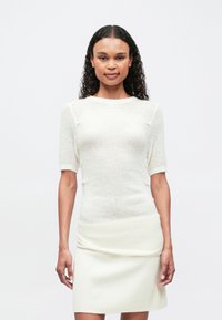 Γυναίκα με σκούρα καστανά σγουρά μαλλιά, ντυμένη με λευκή μπλούζα κοντομάνικη από πλεκτό και αντίστοιχη ριγέ φούστα, που στέκεται μπροστά από ένα απλό ανοιχτό φόντο.