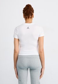Chemise blanche à manches courtes ajustée avec un logo Jordan noir au dos, associée à des leggings gris. Tissu lisse, design simple.