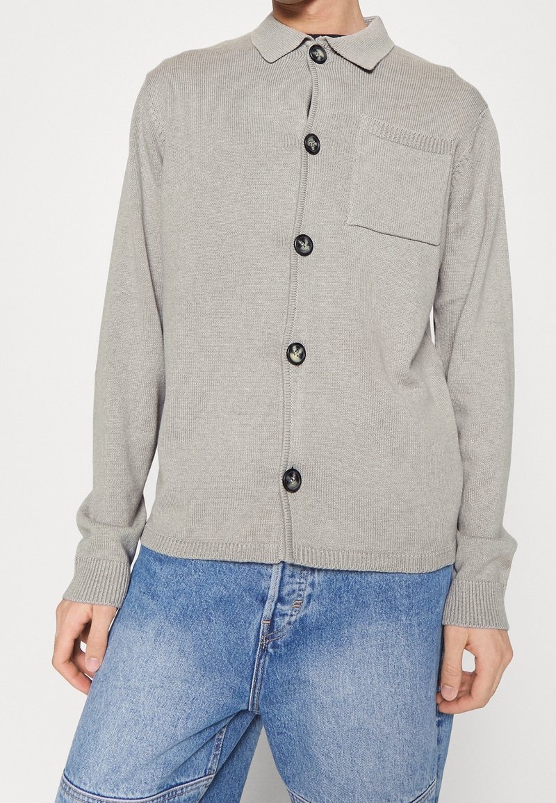 Lichtgrijze gebreide cardigan met een kraag, volledige knopenlijst aan de voorkant en een enkele borstzak. Het heeft ribgebreide manchetten en een ribgebreide zoom.