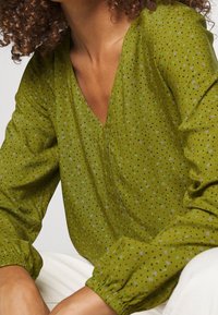 Blusa verde con estampado en forma de V, con pequeños puntos negros, azules y morados, y mangas fruncidas. Textura de tela suave.