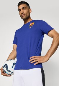 Plava pamučna majica s Barceloninih logotipom i žutim Nike swooshom; nosi se s bijelim šortsevima, držeći šareni lopti za nogomet.