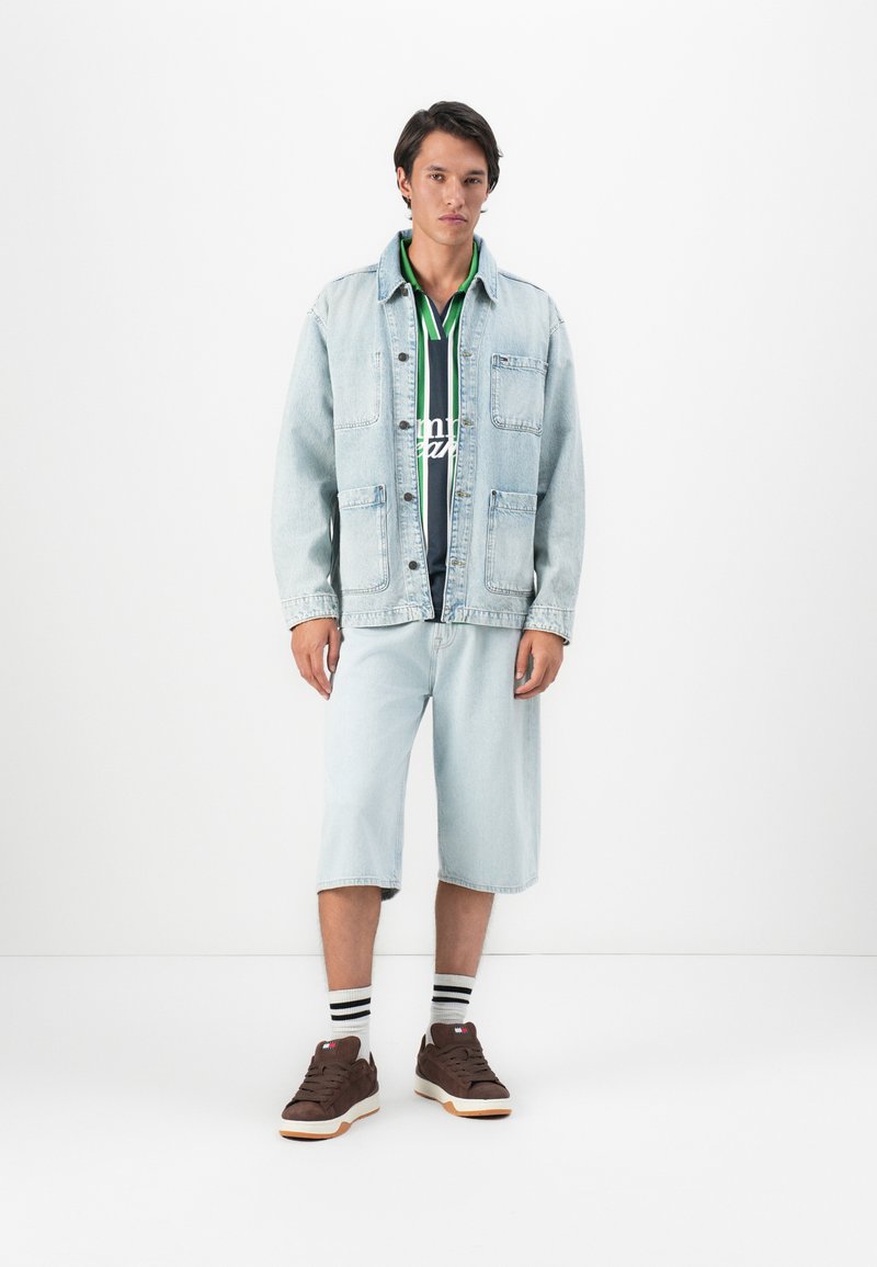 Jeune homme portant une veste en denim clair, un short en denim, un polo rayé vert et bleu marine, des chaussettes rayées et des baskets brunes, se tenant sur un fond blanc.