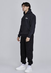 Sudadera navy con bolsillo canguro y logo bordado, combinada con pantalones cargo negros con múltiples bolsillos y zapatillas blancas.