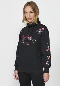 Haut à capuche noir avec une texture en chevrons, broderie florale rouge et bleue sur les manches, capuche à cordon de serrage ajustable et poignets côtelés.