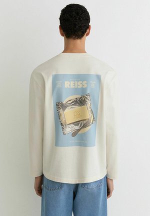 Hombre que lleva una camisa de manga larga color crema con un gráfico de un bloque de mantequilla sobre papel de aluminio y un cuchillo en la espalda, combinado con vaqueros azules.