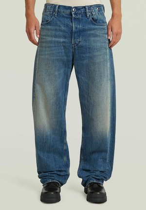 Jean bleu à jambes droites avec des zones décolorées, porté avec des chaussures noires, visible de la taille aux pieds sur un fond neutre.