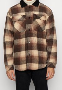 Camicia in flanella a quadri nei toni del marrone e crema, con un collo nero peloso, due tasche sul petto e chiusura anteriore con bottoni. Tessuto testurizzato.