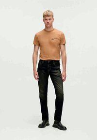 Brūna īsroku T-krekls ar logotipu, saskaņots ar tumšām džinsa biksēm. Modelis valkā melnas ādas kurpes, stāvot pret neitrālu fonu.