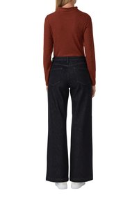 Femme portant un haut à manches longues couleur rouille, un jean large sombre et des baskets blanches, se tenant de dos sur un fond blanc.
