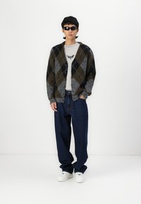 Cardigan lavorato a maglia in un motivo argyle marrone e grigio, indossato sopra una maglietta grigia, abbinato a jeans blu larghi e sneaker bianche.