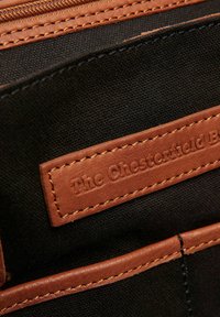 Portafoglio in tela nera con dettagli in pelle marrone, cuciture visibili e un'etichetta impressa che recita "The Chesterfield Brand."