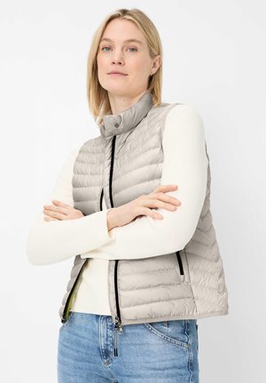 Blonde woman with arms crossed wearing beige quilted vest over white sweater and blue jeans against plain white background.