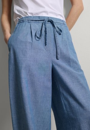 Person trägt lockere hellblaue Leinenhose mit einem vorderen Taillenband und hat die Hände in den Taschen, kombiniert mit einem weißen, in die Hose gesteckten Hemd.