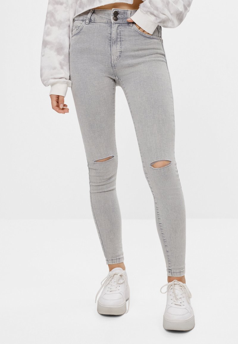 Jeans ajustados en denim gris claro con desgastes en las rodillas y un diseño de cintura alta, combinados con zapatillas blancas.