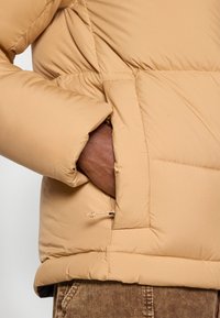 Burgundska puffer jakna s kvadratnim dizajnom, koja ima bočnu džepu i podesivu vezicu na rubu za prilagodbu kroja.