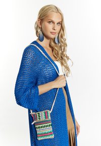 Modrý pletený cardigan s širokými rukávmi, prehodený cez hnedý outfit, držiaci viacfarebnú tkanú tašku s geometrickými vzormi.