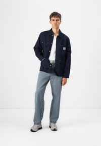 Mørkeblå denim skjortejakke med to brystlommer, sølvknapper og et logomerke. Samtidig med lyseblå jeans og grå sneakers.