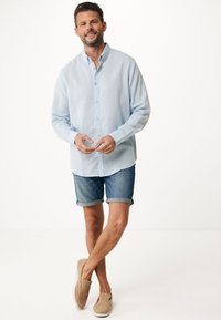 Camisa de botones azul claro hecha de lino, combinada con pantalones cortos de mezclilla enrollados. El modelo lleva zapatos de lona beige.