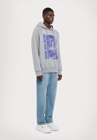 Felpa grigia con stampa grafica in viola. Tessuto morbido, cappuccio regolabile con lacci, vestibilità rilassata. Abbinata a jeans azzurri e sneakers bianche.