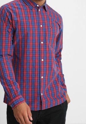 Chemise à carreaux rouges et bleus, à manches longues et boutons blancs. Tissu en coton avec un col standard et un logo visible.