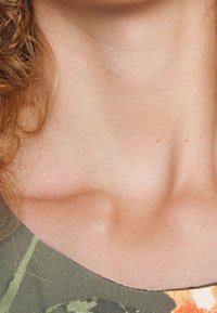 Zielony materiał w kamuflażu na dekolcie o subtelnej teksturze prążkowanej. Gładka skóra w kolorze ciała widoczna poniżej, podkreślająca obszar obojczyków. Brak wyraźnych wzorów.