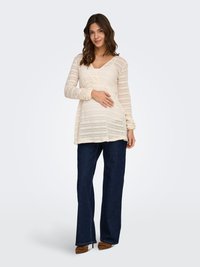 Ženska z dolgimi rjavimi lasmi, oblečena v kremasto pleteno pulover in temno modre kavbojke, se smehlja z eno roko, ki počiva na njenem trebuhu.