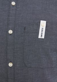 Camisa de botones gris oscuro con una tela texturizada, que presenta un bolsillo en el pecho y una etiqueta blanca marcada "COMFORT STRETCH". Los botones son transparentes.