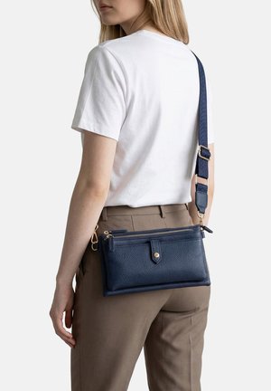 Donna con maglietta bianca e pantaloni marroni che porta una borsa a tracolla in pelle blu navy con tracolla larga regolabile sulla spalla.