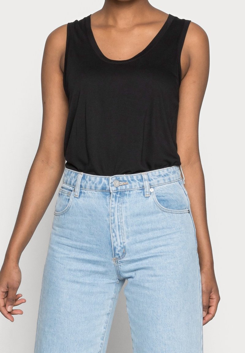 Personne portant un haut noir sans manches rentrée dans un jean en denim bleu clair taille haute avec des poches avant et une fermeture par bouton.