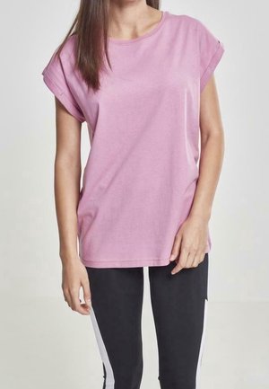 Kobieta ubrana w różowy t-shirt z krótkim rękawem oraz czarne legginsy z białymi paskami po bokach, stoi na tle gładkiego tła.