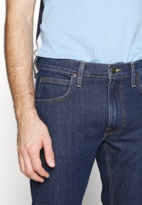 Tmavě modré džínové kalhoty s designem pěti kapes, žlut ým šitím a kovovým knoflíkem. Nošeny s světle modrou košilí.