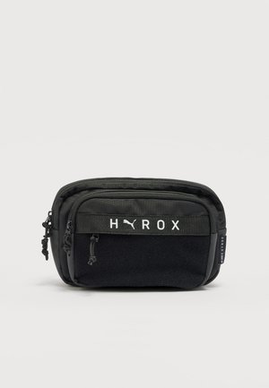 Čierna obdĺžniková ľadvinka s viacerými priehradkami na zips a logami "HYROX" a Puma na prednom paneli.