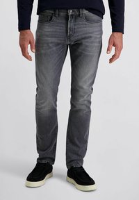 Homme portant un jean droit gris délavé et des chaussures noires à enfiler avec des semelles blanches, se tenant devant un fond uni.