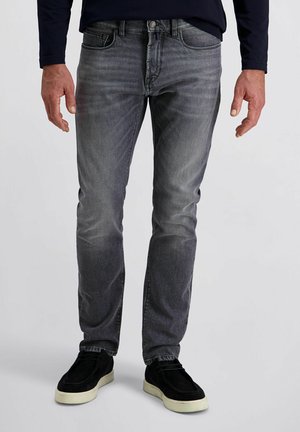 Mann in ausgewaschenen grauen Straight-Leg-Jeans und schwarzen Slipschuhen mit weißen Sohlen, der vor einem schlichten Hintergrund steht.