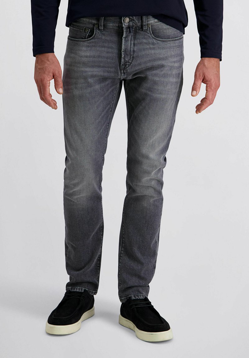 Homme portant un jean droit gris délavé et des chaussures noires à enfiler avec des semelles blanches, se tenant devant un fond uni.