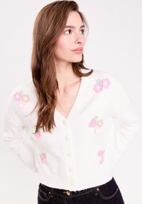 Gilet blanc avec un col en V et une fermeture à bouton, ornée d'embellissements floraux en rose, beige et vert. Texture douce ; design court.