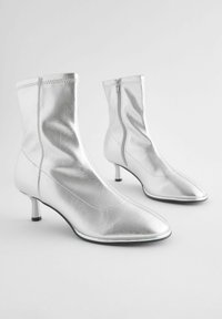 Srebrne čizme do gležnja s elegantnim dizajnom, špicastim vrhom i potpeticom srednje visine. Napravljen od sjajnog metalnog materijala s bočnim patentnim zatvaračem.
