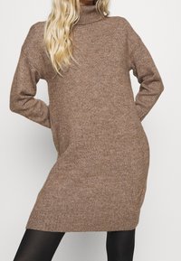 Robe en tricot à col montant de couleur marron clair avec une silhouette décontractée, des poignets côtelés et une texture douce, se terminant au-dessus des genoux.