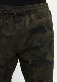 Kamotové tepláky ve tmavě zelené barvě, vyrobené z texturované tkaniny, s elastickým pasem a šňůrkou na stažení. Výrazný maskáčový vzor zvyšuje vizuální atraktivitu.