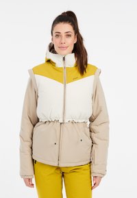 Pūkuotas švarkas su šviesiai ruda apatine dalimi ir balta viršutine dalimi, turintis geltoną kepuraitę, raišteliu reguliuojamą liemenį ir užtrauktuku užsegamą priekinę dalį.