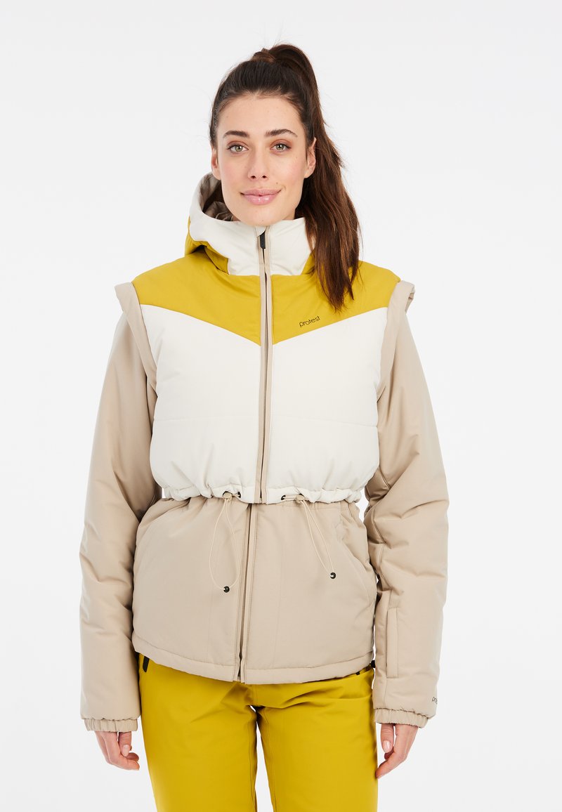 Pūkuotas švarkas su šviesiai ruda apatine dalimi ir balta viršutine dalimi, turintis geltoną kepuraitę, raišteliu reguliuojamą liemenį ir užtrauktuku užsegamą priekinę dalį.