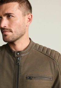 Braune Lederjacke mit Reißverschluss, Druckknopfkragen und strukturierten Schulterakzenten. Enthält eine mit Reißverschluss verschließbare Brusttasche.