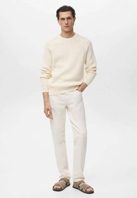 Suéter de punto cable en crema, pantalones blancos y sandalias marrones. El suéter tiene un escote redondo y un patrón texturizado; los pantalones son de pierna recta.