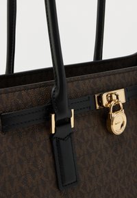 Καφέ υφασμάτινη τσάντα με μαύρες δερμάτινες λαβές, χρυσές μεταλλικές λεπτομέρειες και κλειδαριά στο μπροστινό μέρος. Γεωμετρικό μοτίβο στο υλικό.