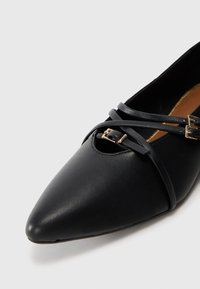 Ballerine in pelle nera con punta affusolata, dotate di nastri incrociati e un piccolo dettaglio in metallo a forma di fibbia. Texture liscia in tutto il modello.