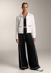 Fehér, zipzáros kabát fekete részletekkel, laza fekete nadrággal, fehér oldalsávokkal párosítva. Sima textúra, laza szabás és sportos dizájn.