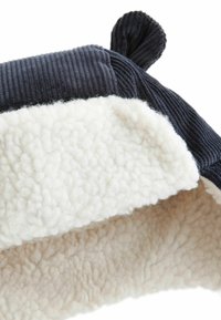 Tmavomodrá kordurová čiapka s bielou fleecovou podšívkou, s okrúhlym tvarom a malým detailom na prednej strane. Mäkko, textúrované materiály.