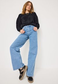 Black fuzzy sweater, light blue wide-leg denim jeans, and black sneakers with textured accents and a ribbed sole. Model poses with arms on hips.