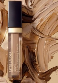 Estee Lauder Futurist concealer i en klar tube med en svart hette og gull aksenter, omkranset av flekker av forskjellige brune nyanser.